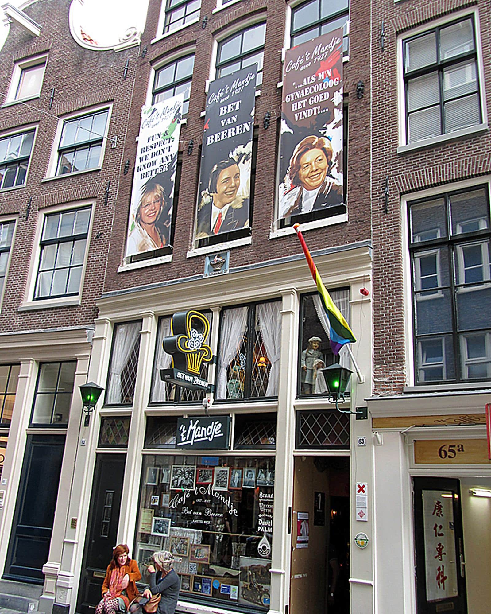 De gevel van café ’t Mandje op de Zeedijk, met vaandels waarop Bet van Beeren lachend in pak staat, en twee vrouwen op het bankje voor de deur