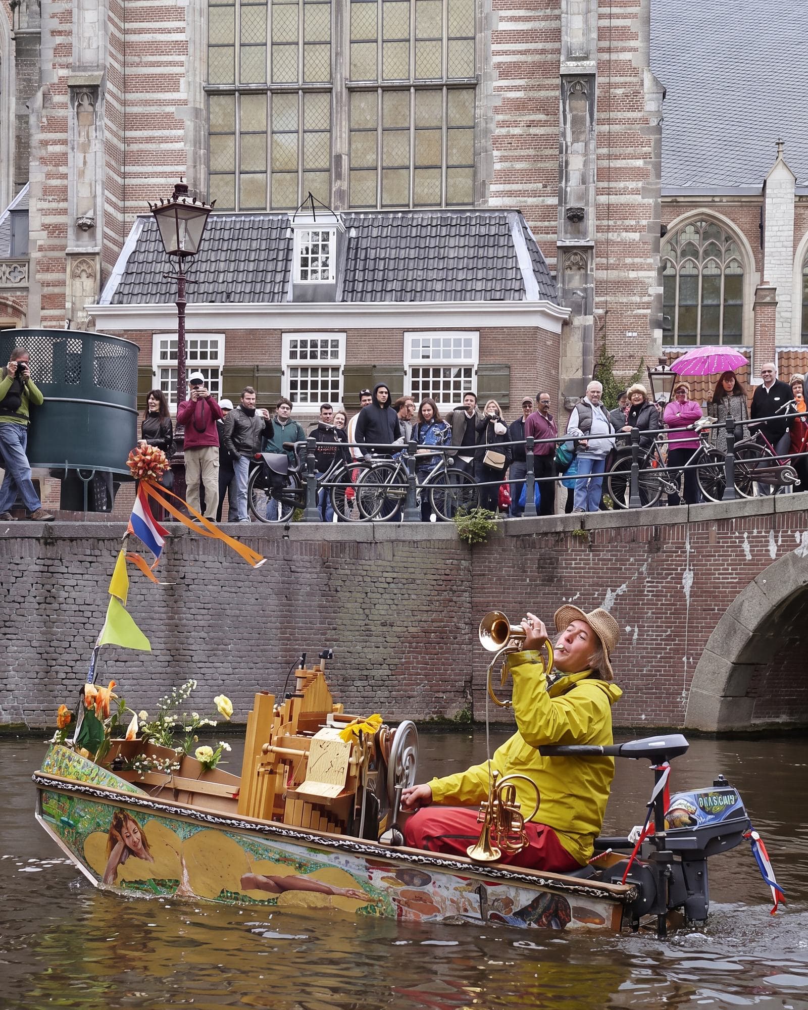 Reinier Sijpkens in een geel regenpak speelt trompet in zijn bootje De Notendop, met bloemen en een orgeltje aan boord, onder een brug vol publiek